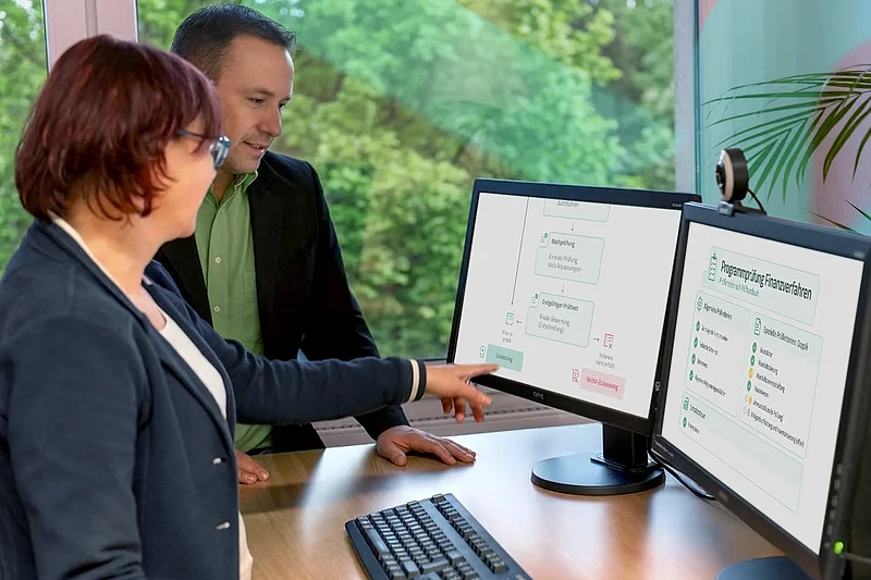 Eine Mitarbeiterin und ein Mitarbeiter prüfen an zwei Monitoren den Ablauf einer Programmprüfung für Finanzverfahren.